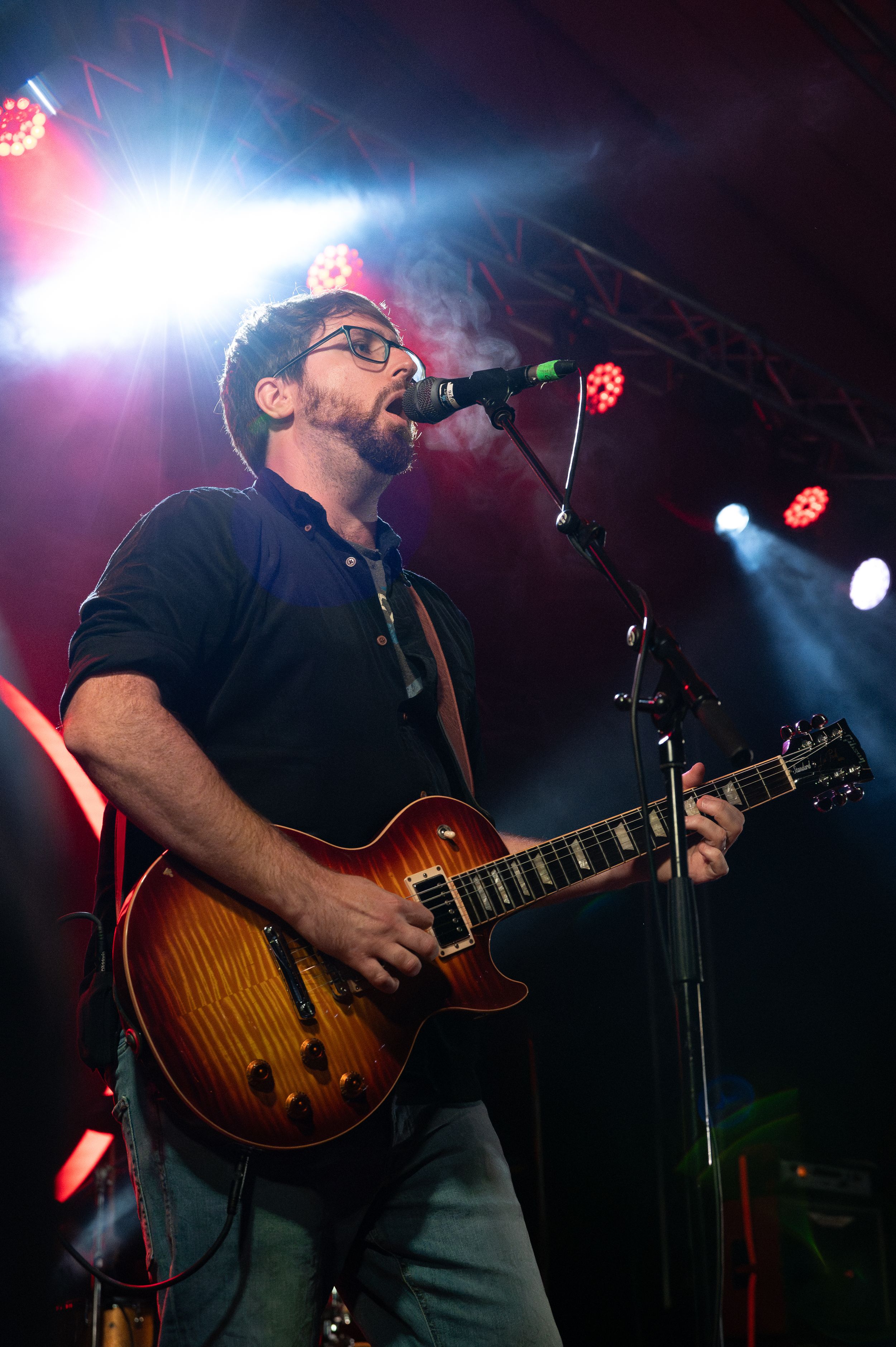 Guitarist performs with sunburst Les Paul guitar under colorful stage lighting.