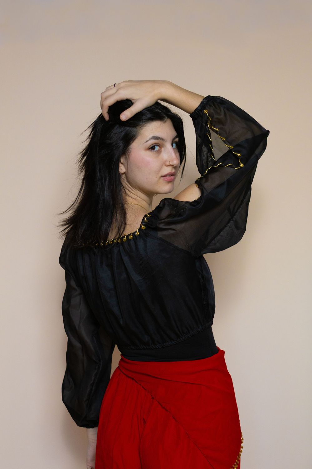 Fashion portrait of model in black blouse and red skirt against beige wall.