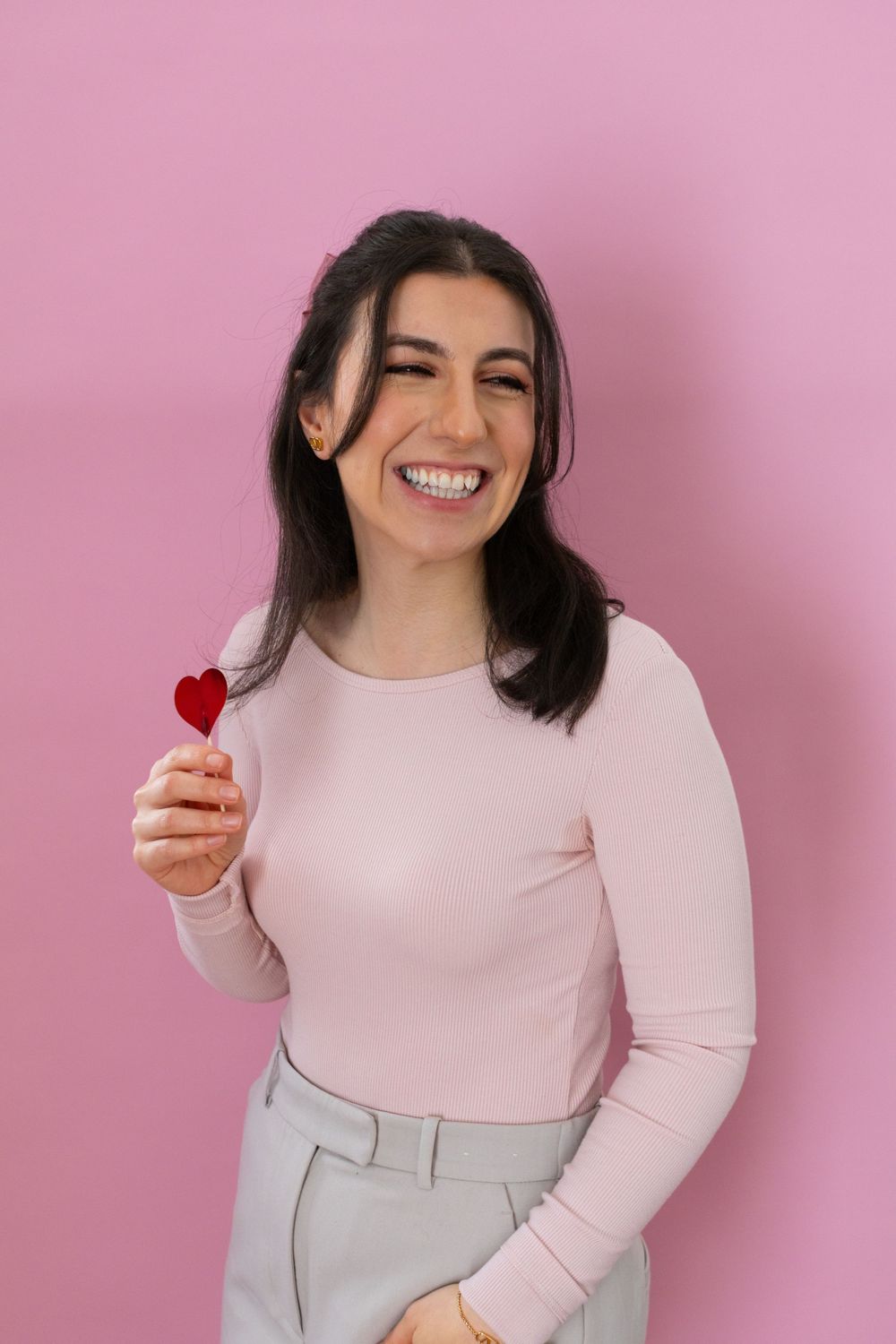 A person in a pink sweater poses against a pink background holding a heart-shaped object in a playful series of photos.