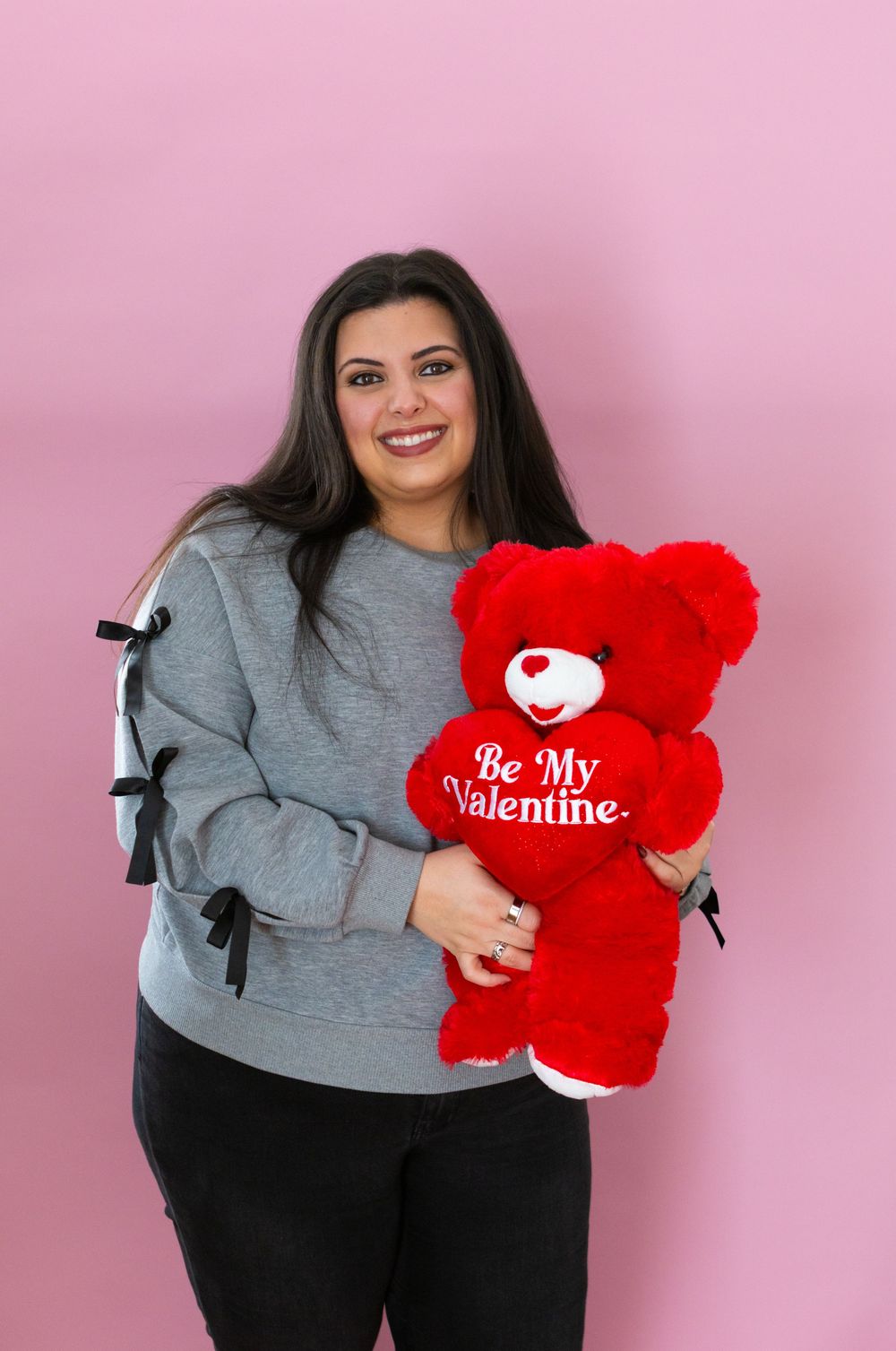 Woman in gray sweater holds red 'Be My Valentine' teddy bear against pink background in Valentine's Day photo series.