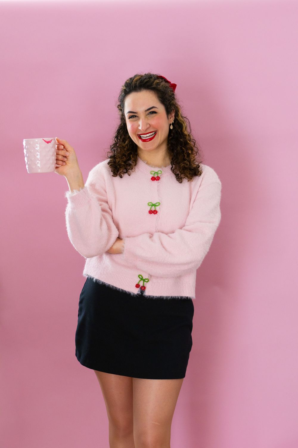 Someone wearing a pink sweater with cherry embroidery and black mini skirt holds a polka dot mug against pink background.
