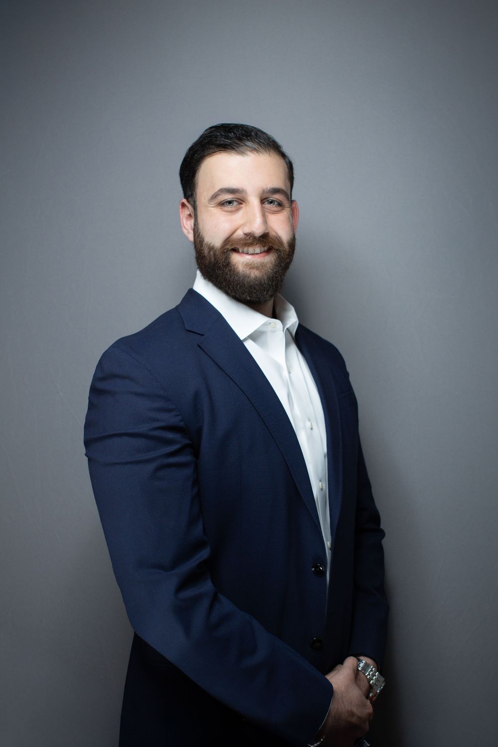 Smiling bearded man in navy blazer and white shirt poses confidently against a gray background.