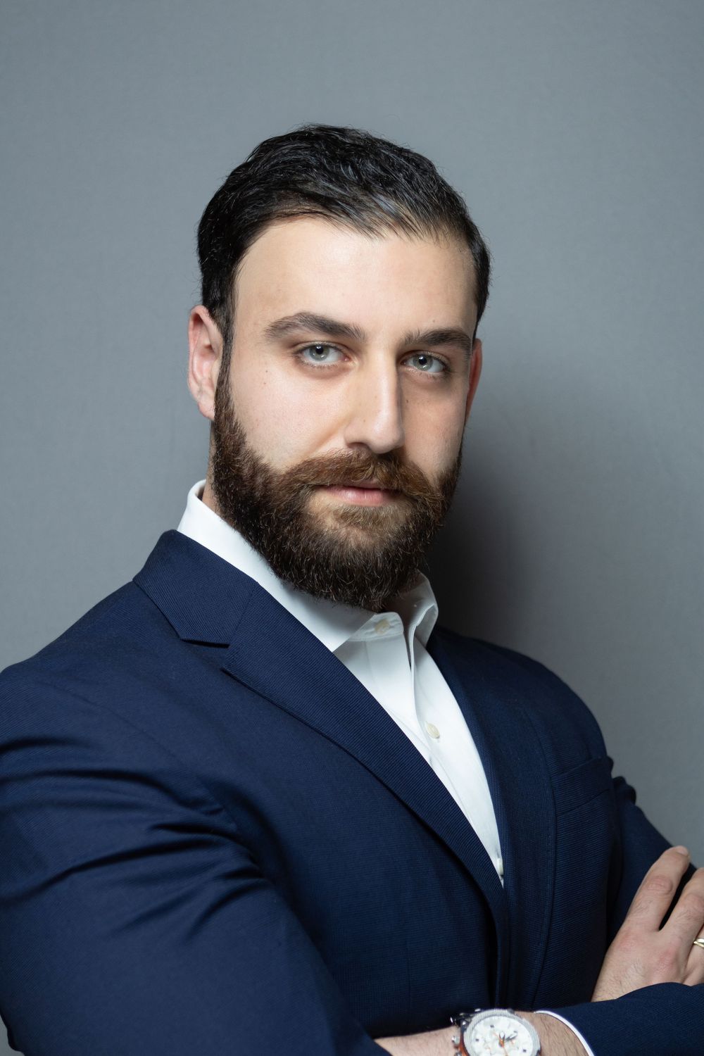 Professional man with beard in navy suit, arms crossed, posing confidently against gray background.