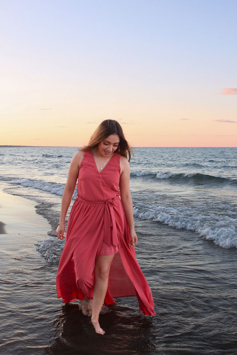 Woman in flowing red dress walking through ocean waves at sunset.