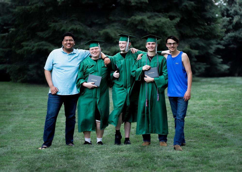 College graduates in green graduation gowns pose together on a grassy lawn for a casual group photo session.