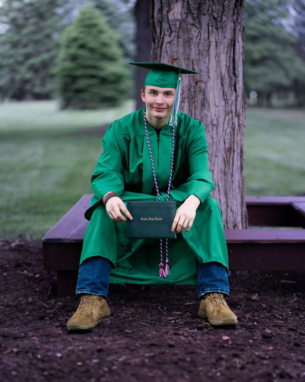 Graduate in emerald green cap and gown sits on wooden bench by tree trunk holding diploma during graduation photoshoot.