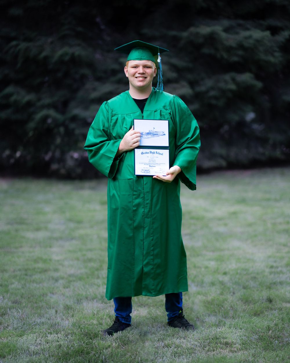 Graduate in emerald green cap and gown holds diploma while standing on grass outdoors.