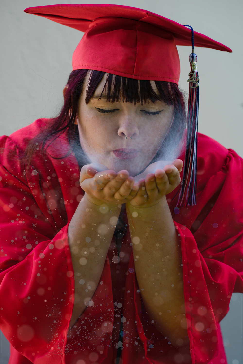 A graduate in red cap and gown blows glittery sparkles from cupped hands in a celebratory graduation portrait.