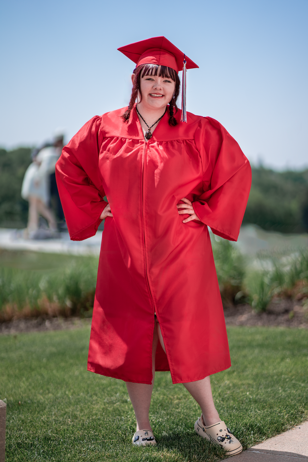 Young graduate in bright red cap and gown poses playfully on green grass on graduation day.