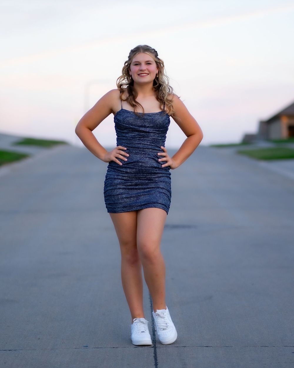 A person in a sparkling navy blue dress and white sneakers poses confidently on a suburban street at dusk.