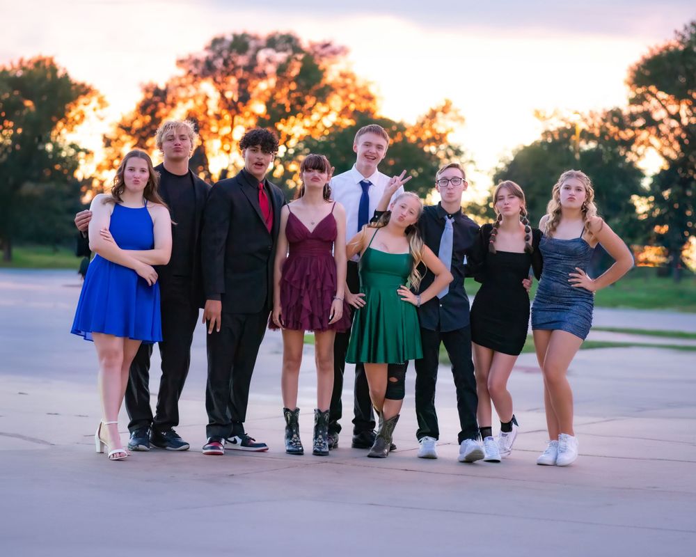 A group of formally dressed young people pose together outdoors against a sunset sky.