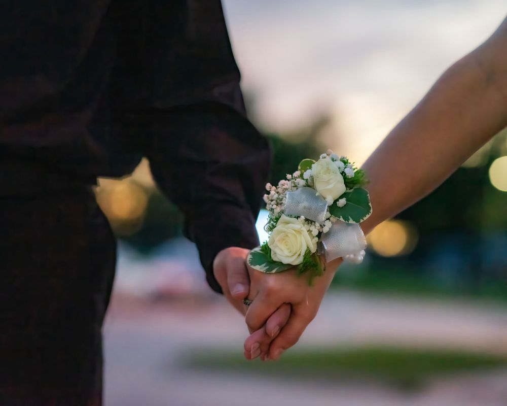 A couple holding hands with a white and green floral corsage wristlet against a soft bokeh background.