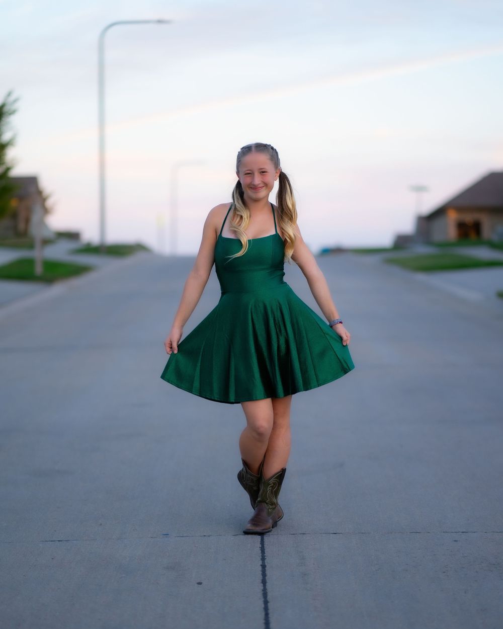 A person in a green dress and brown cowboy boots spins and twirls on a concrete path at sunset.