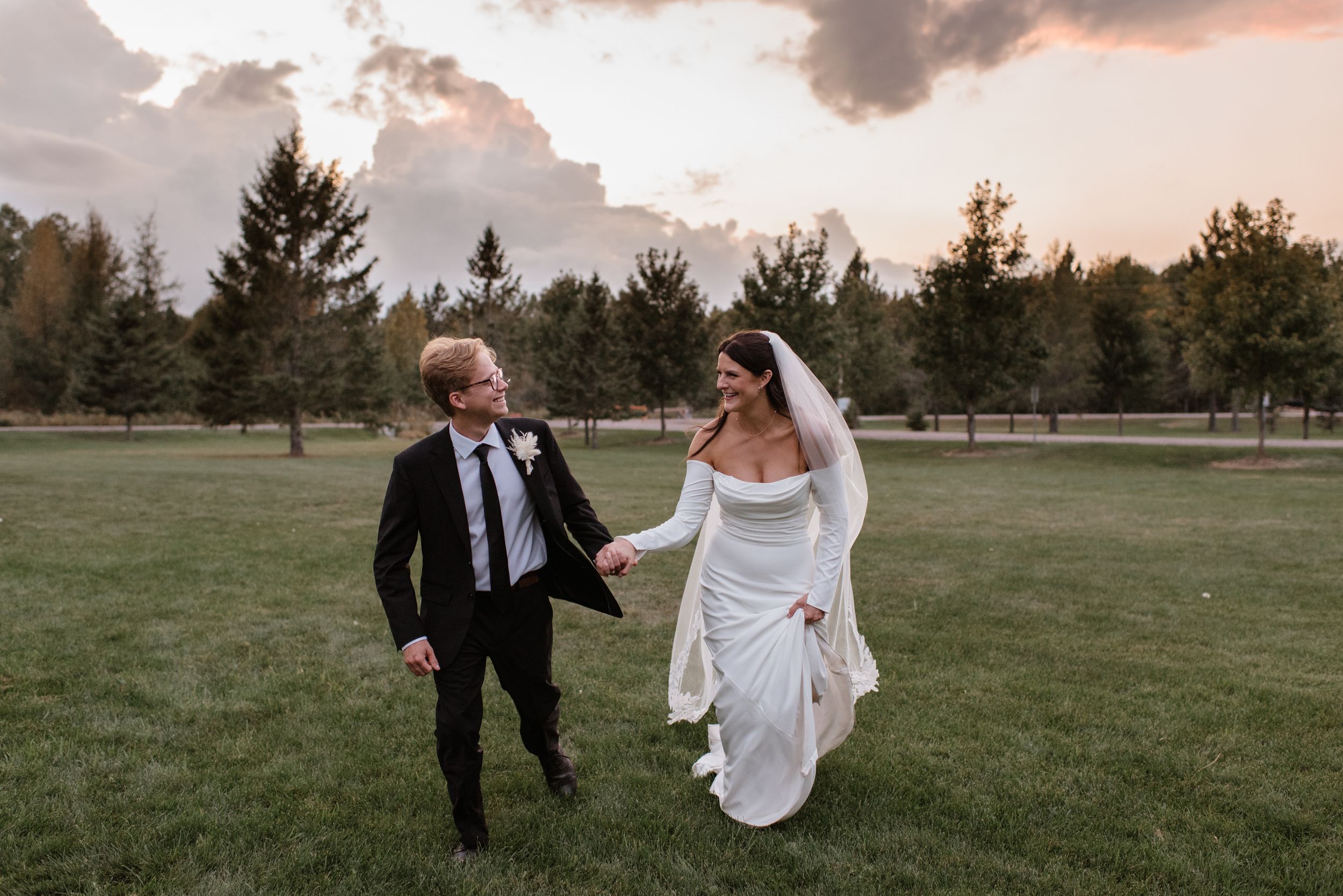 Bride and groom walking hand in hand on green lawn at sunset with pine trees and dramatic sky backdrop.