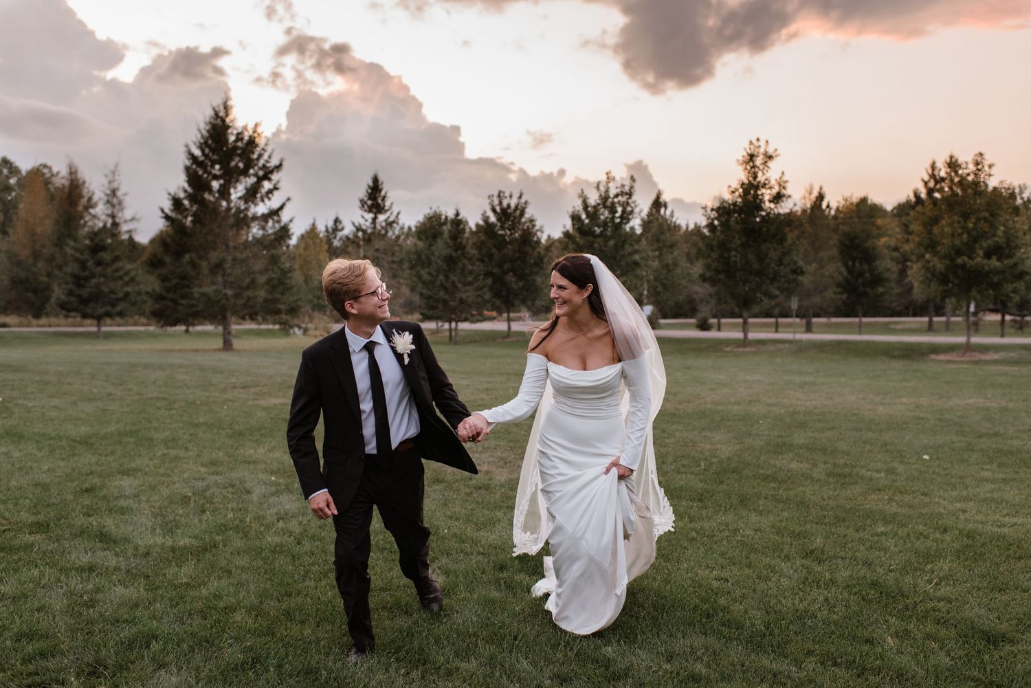 Bride and groom walking hand in hand on green lawn at sunset with pine trees and dramatic sky backdrop.