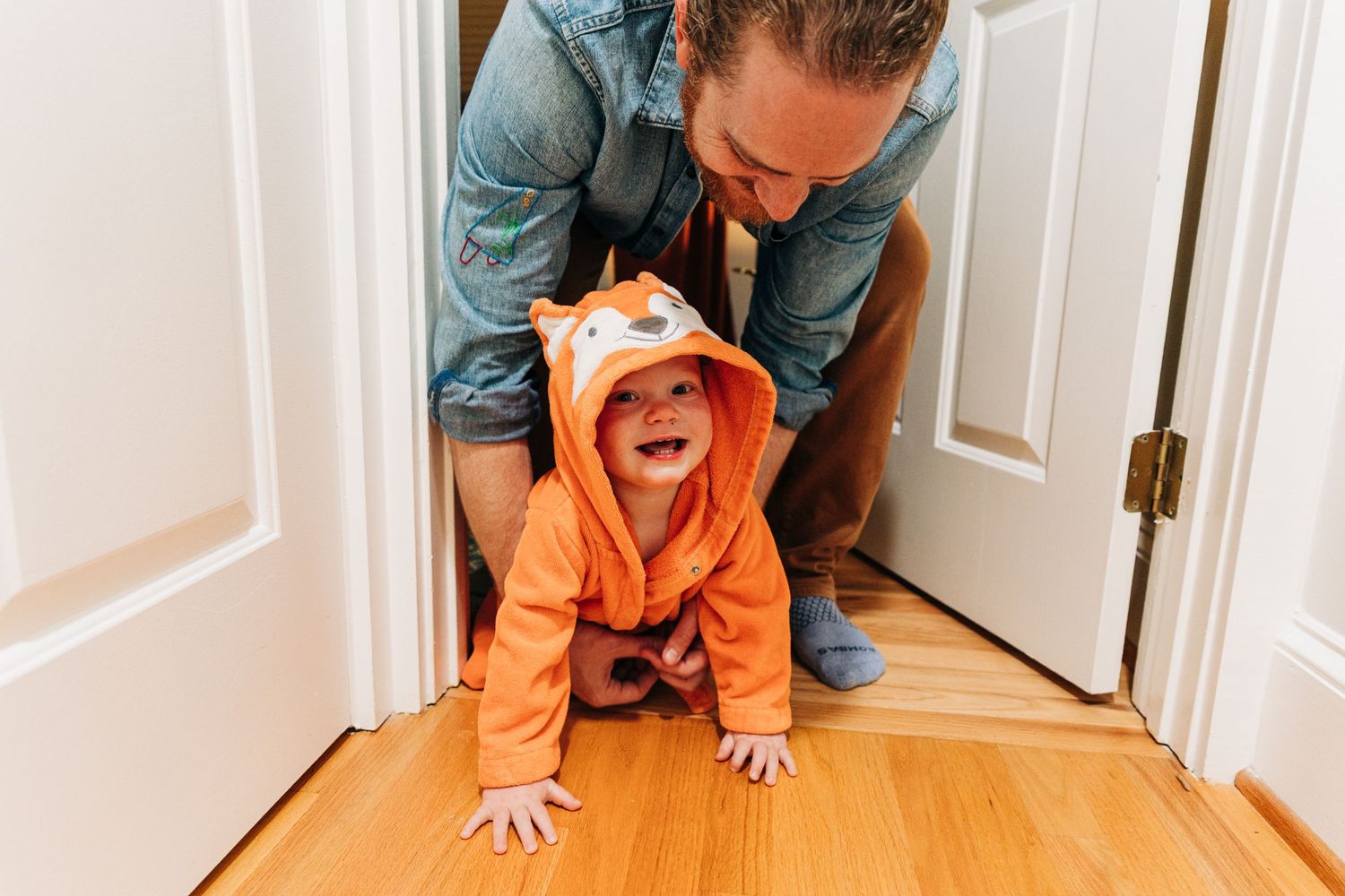 A laughing baby in an orange fox towel crawls on hardwood floors while an adult leans over them in a doorway.