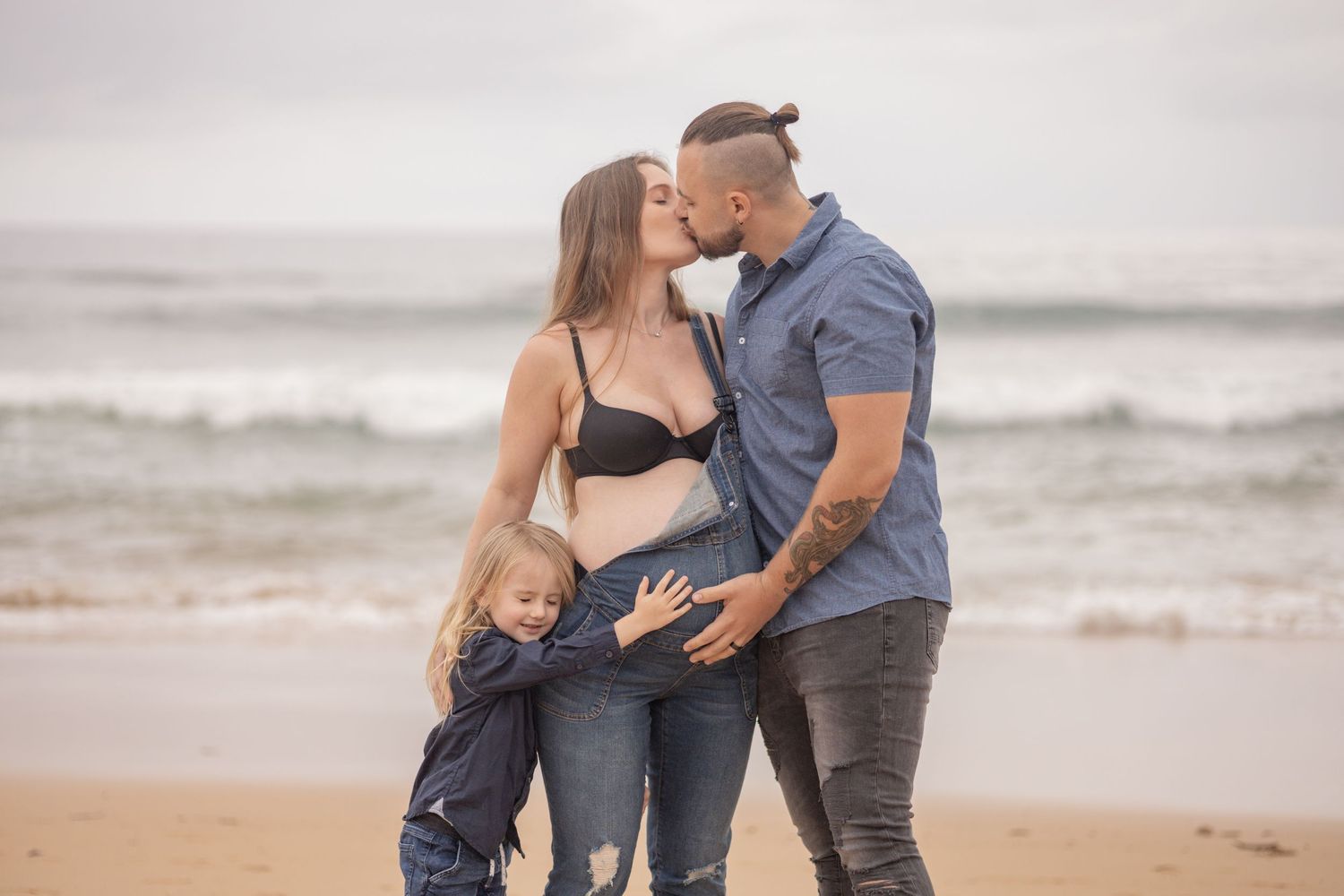 Couple kissing on beach with young child between them, pregnant woman showing baby bump.