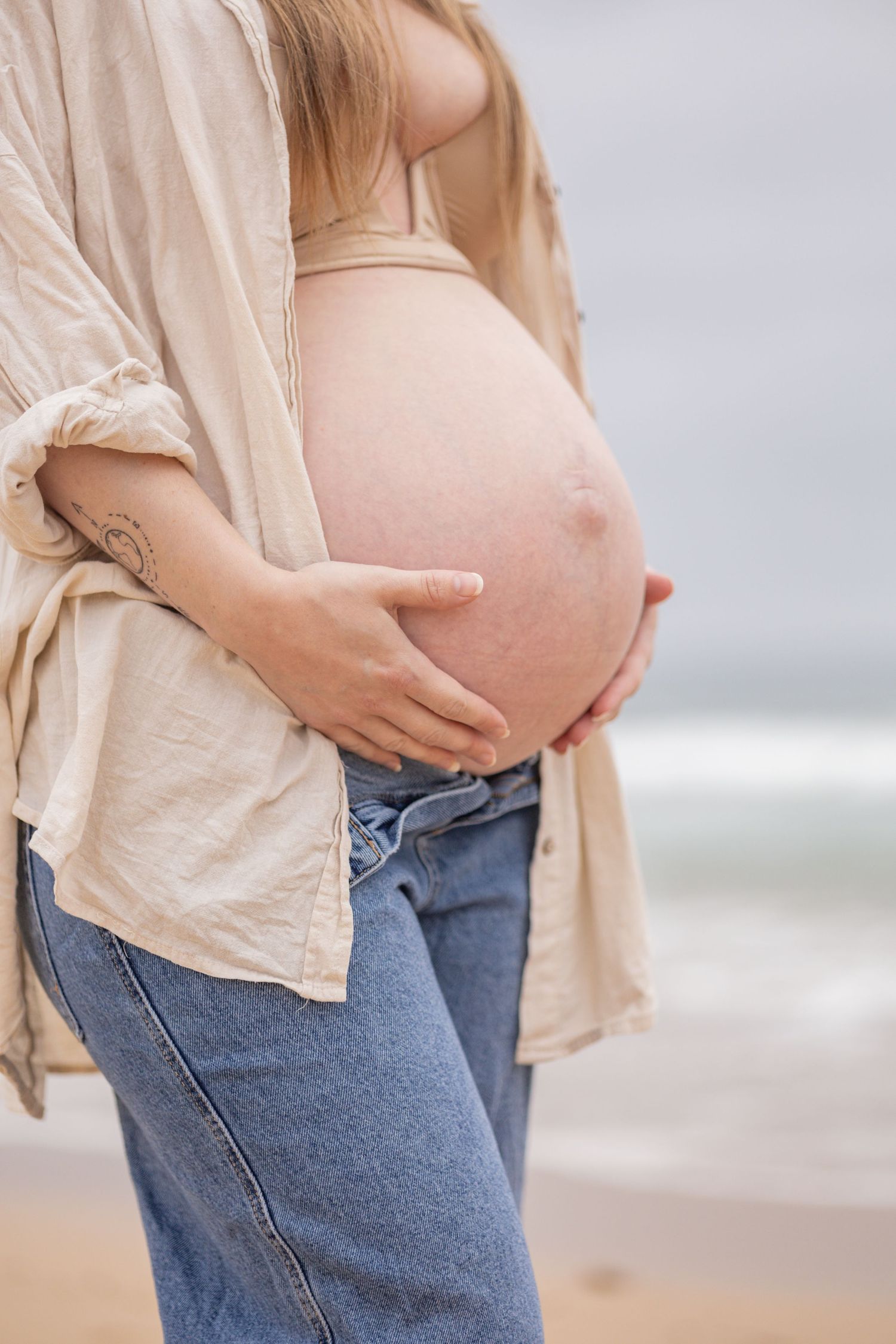 Close-up of pregnant woman in beige shirt and jeans cradling bare baby bump at beach.