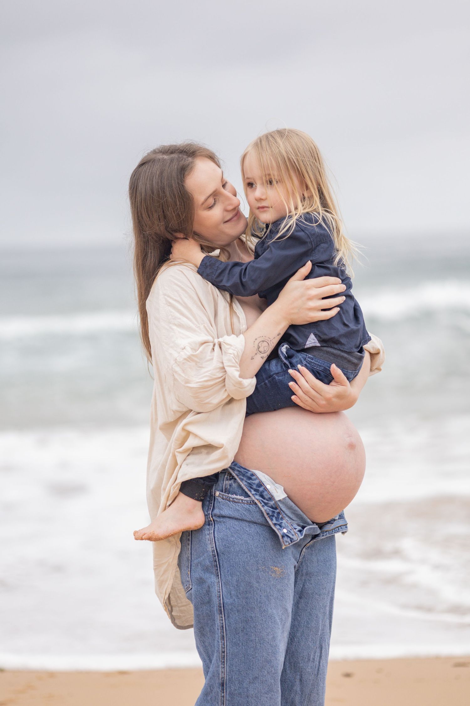 Pregnant woman in beige shirt and jeans lovingly holds young blonde child on beach by ocean.