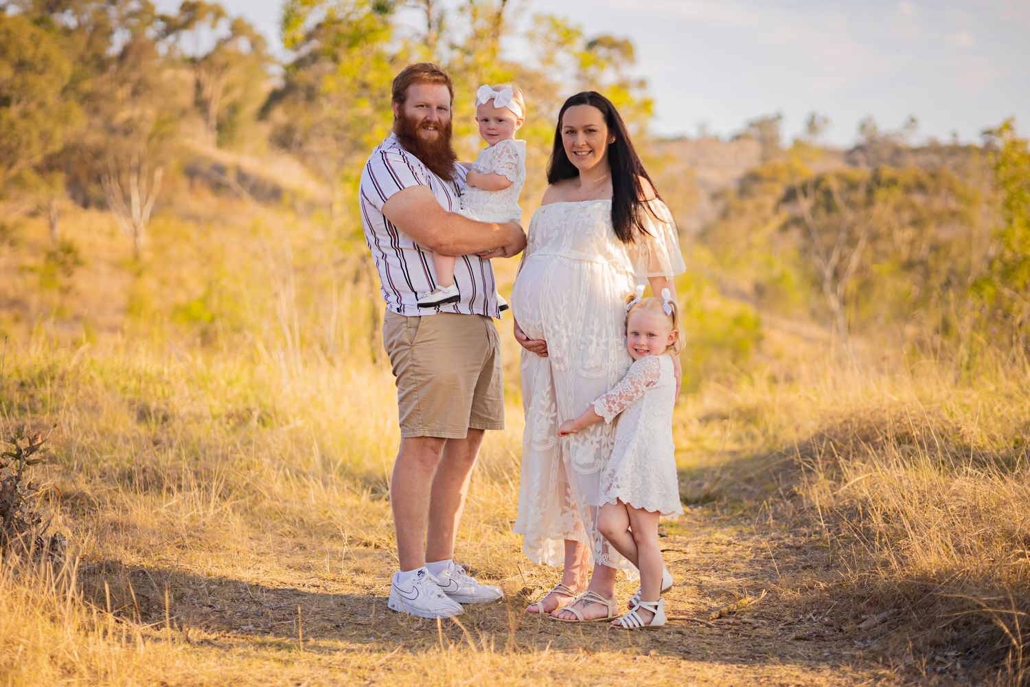 Happy family with bearded dad, pregnant mom, and two young children posing in golden field outdoors.
