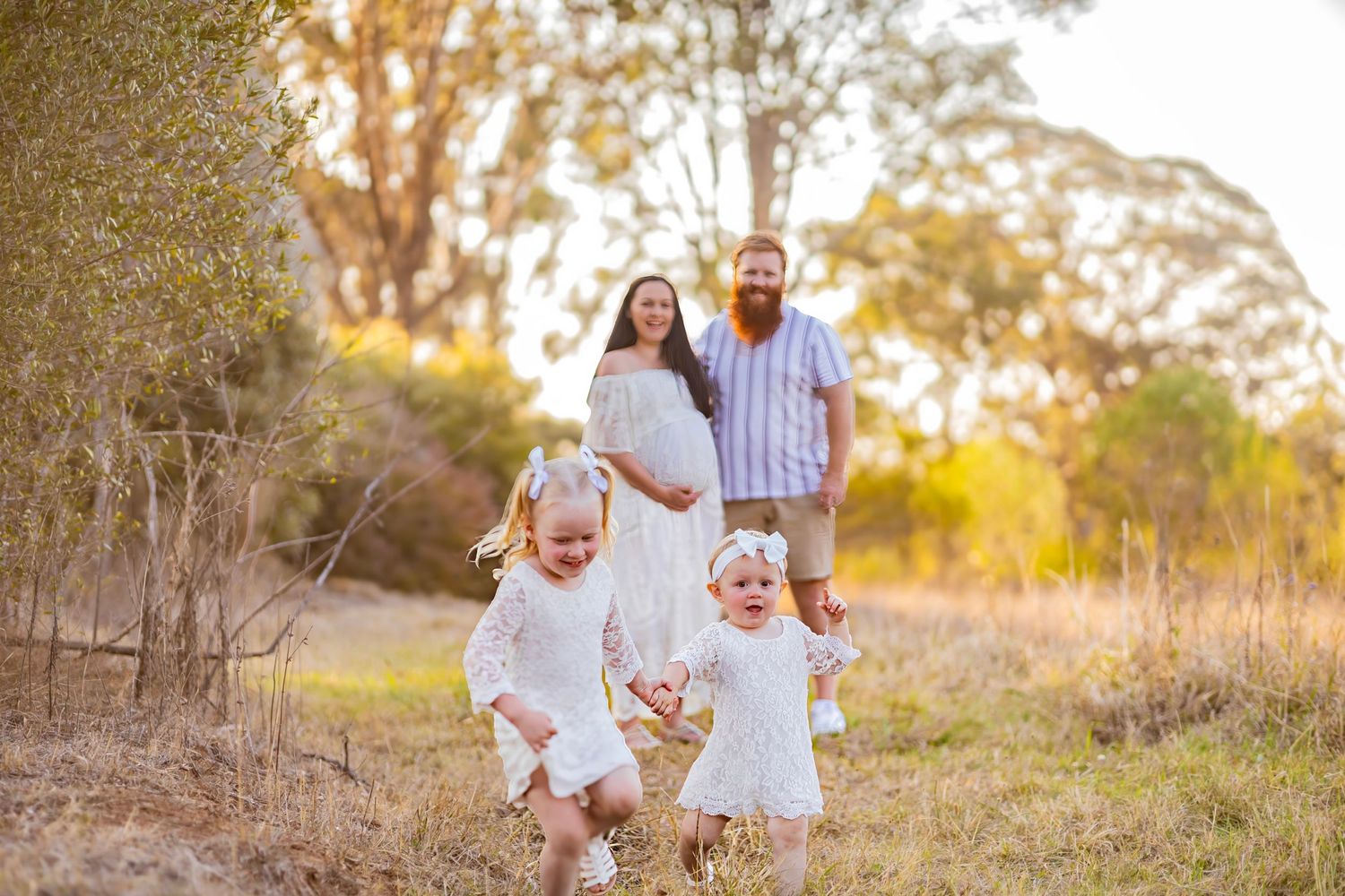 Happy family with pregnant mother and twin toddler girls in white dresses walk in golden field.
