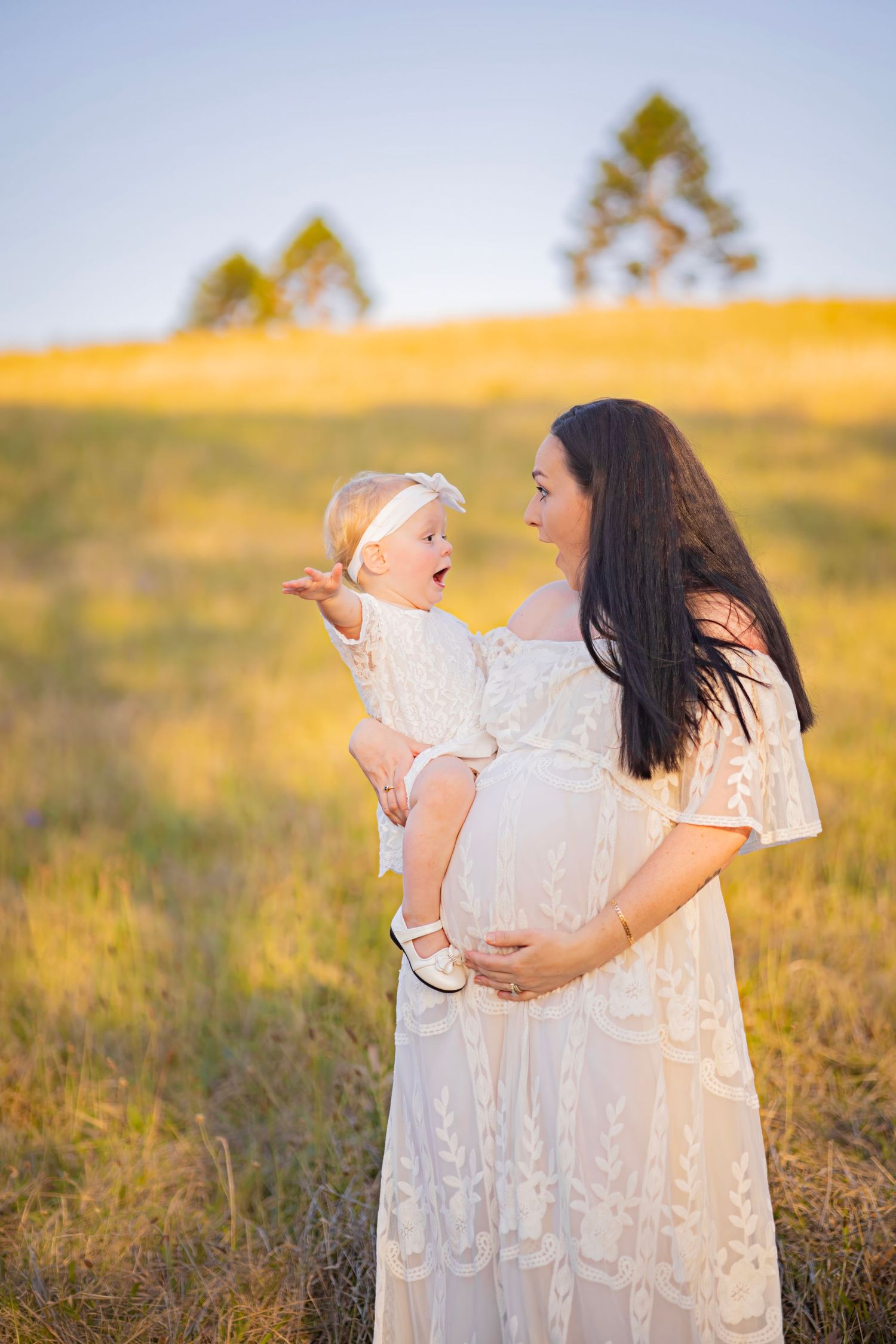 Pregnant woman in white lace dress holding baby girl in sunlit meadow during golden hour.