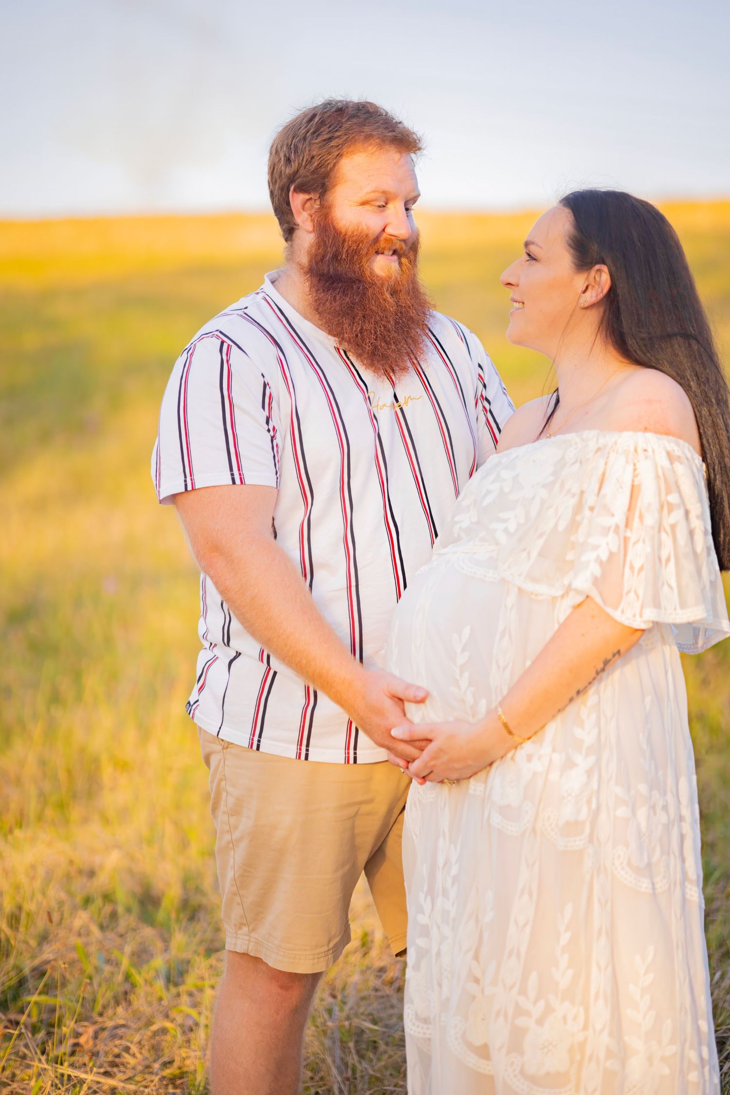 Bearded man and pregnant woman in white dress holding hands smiling at each other in sunny field.