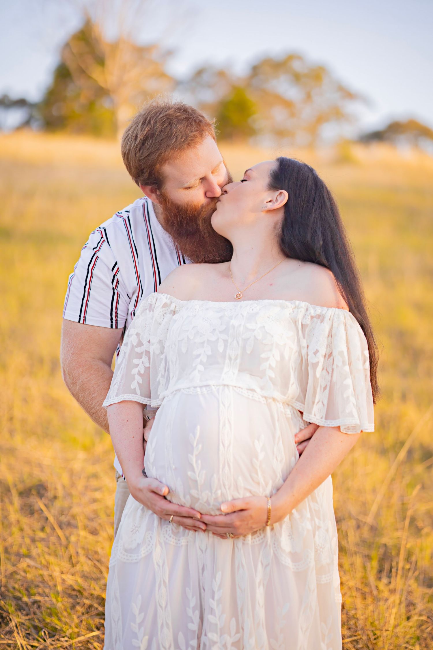 Couple kissing in golden field, man embracing pregnant woman in white lace off-shoulder dress at sunset.