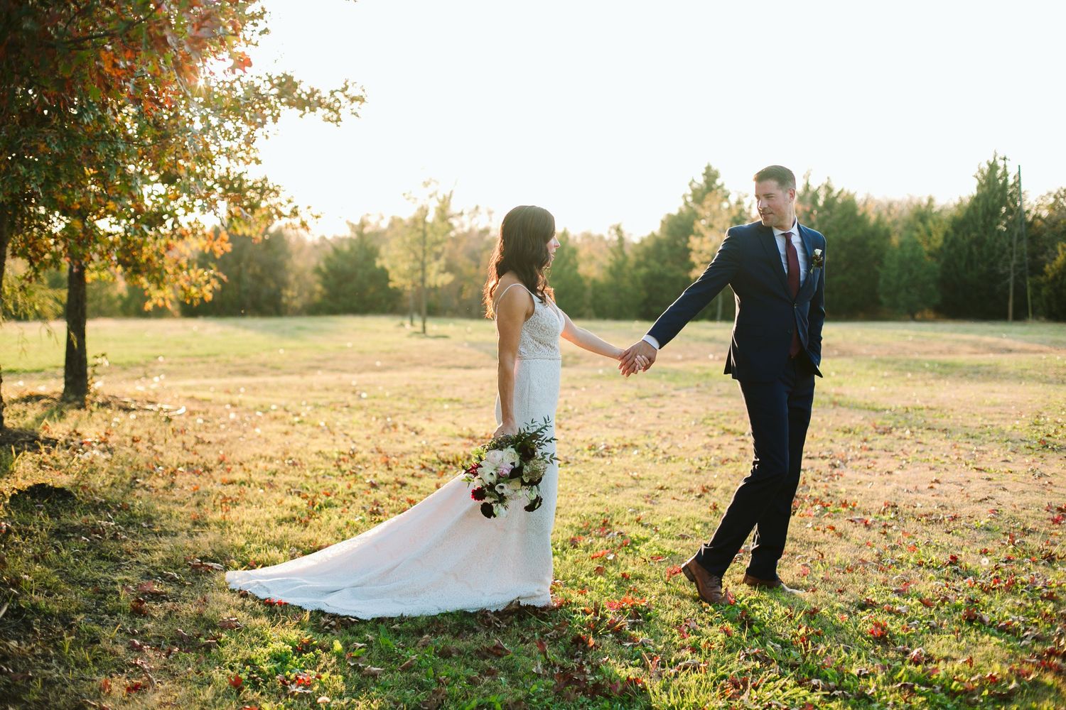 Bride and groom holding hands in a sunlit autumn field, bride wearing a white gown with a floral bouquet.