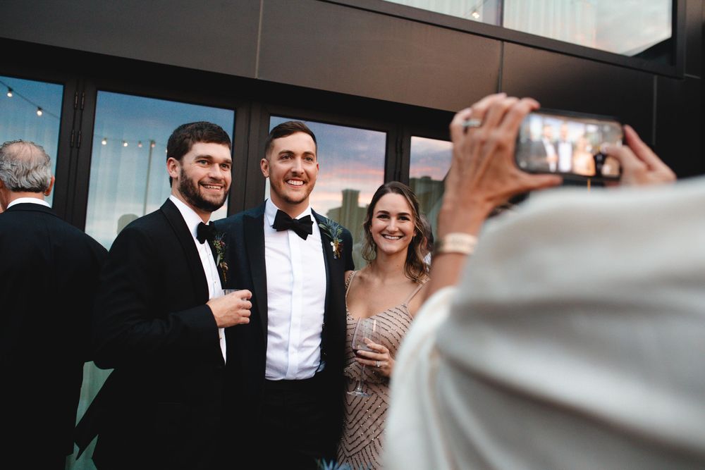 Two men in tuxedos and a woman in a sequin dress pose for a photo at a formal wedding reception.