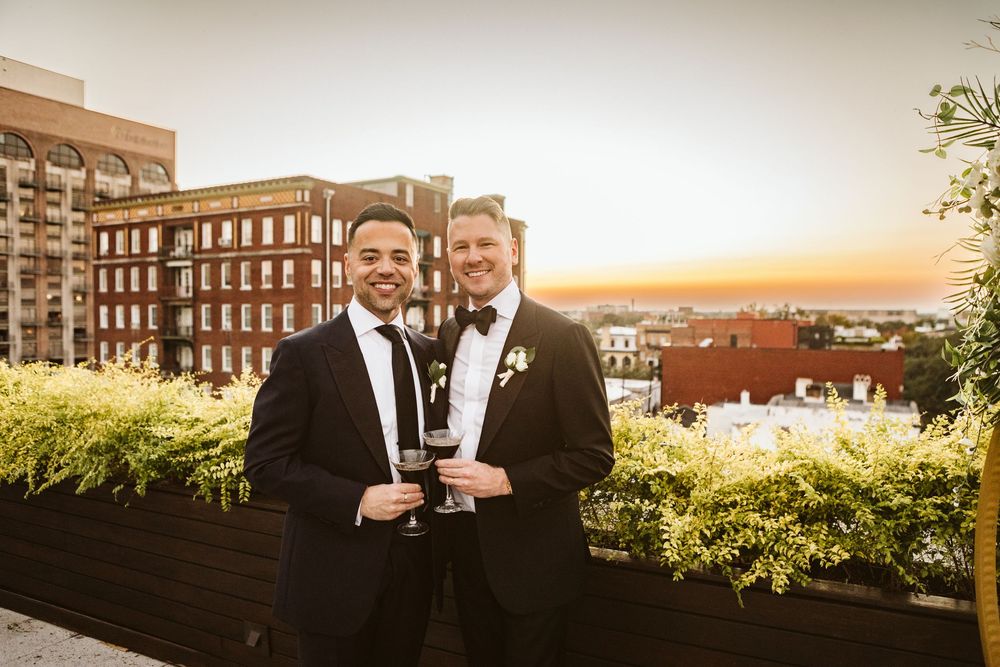 Two grooms in tuxedos toast with wine glasses on a rooftop at sunset with city skyline views.