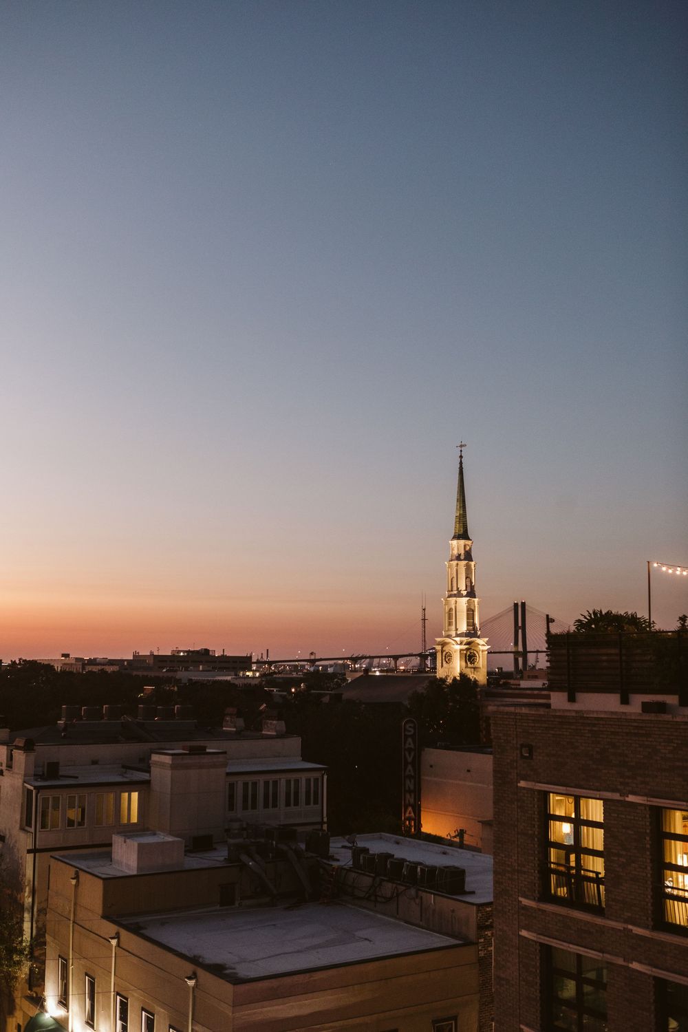 Twilight cityscape with illuminated church steeple rising above rooftops against a gradient sunset sky.