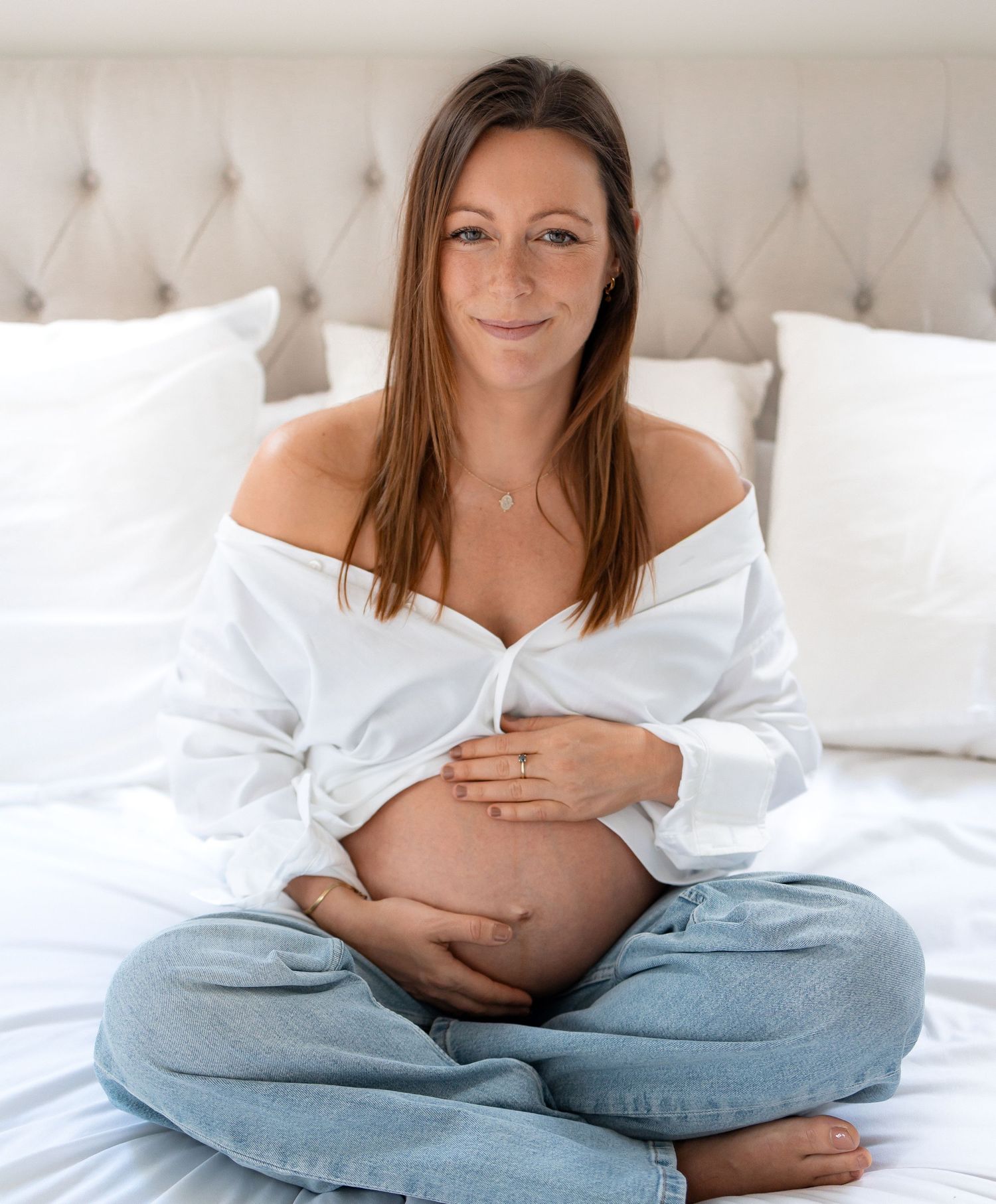 Pregnant woman sitting cross-legged on white bed, cradling bare belly, wearing open white shirt and jeans.