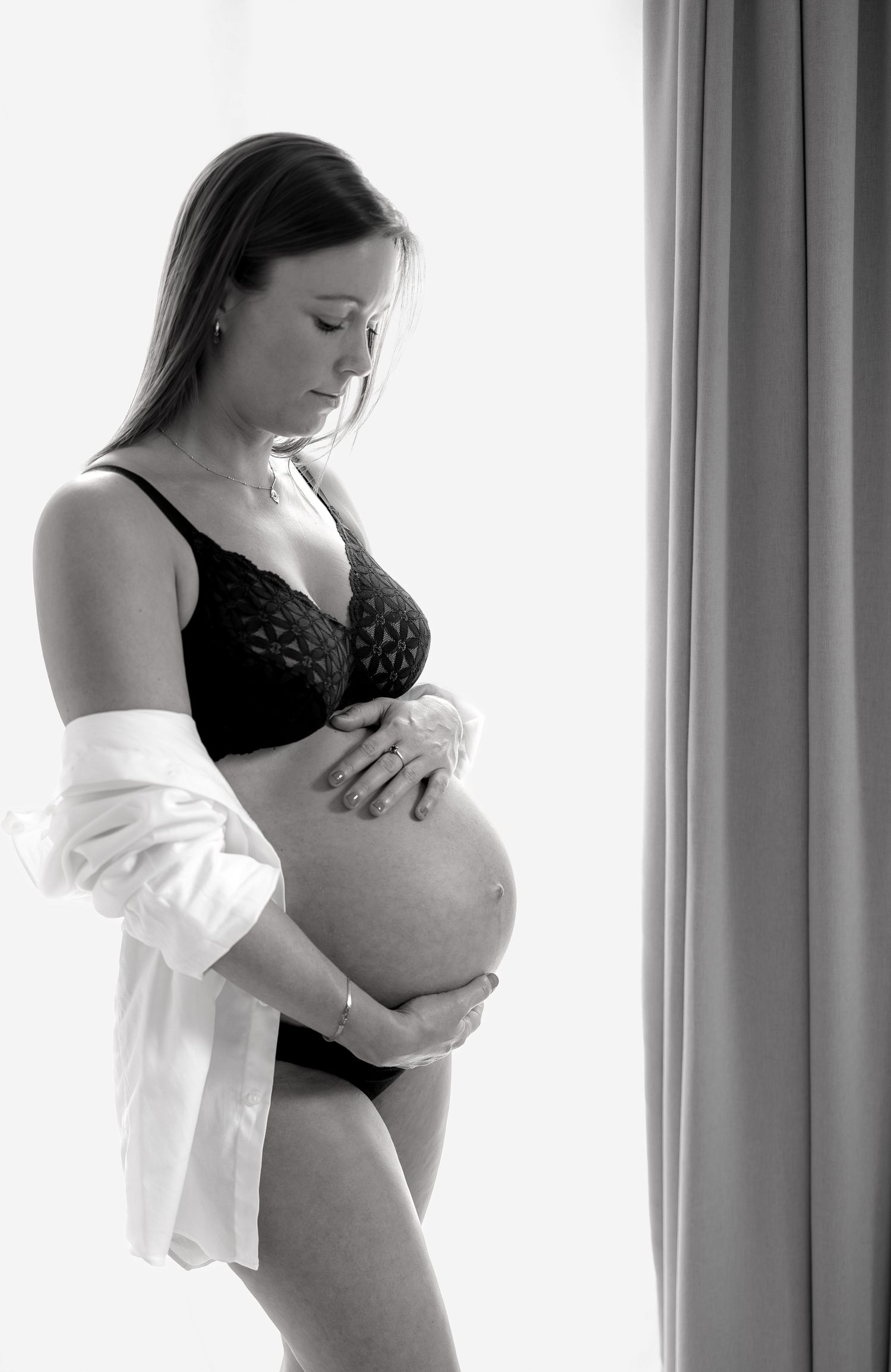 Black and white photo of a pregnant woman in lingerie cradling her baby bump near a bright window.