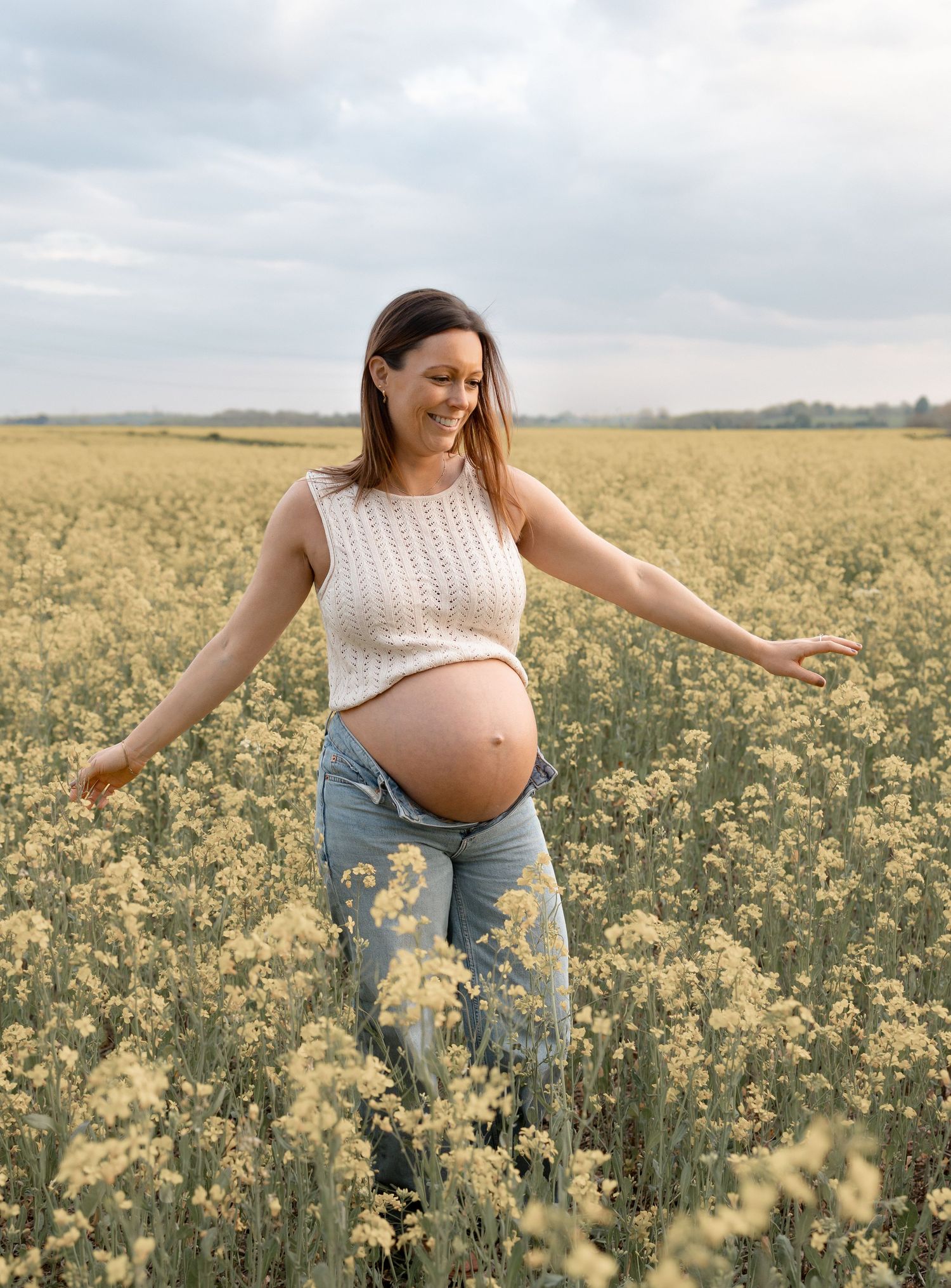 Smiling pregnant woman in white top and jeans standing in a blooming yellow flower field with arms outstretched.