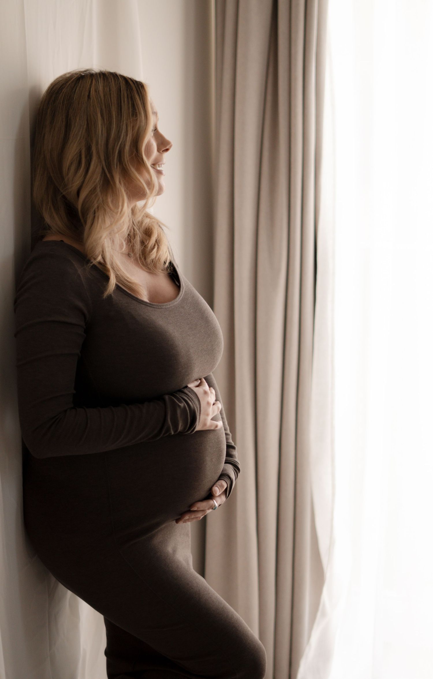 Pregnant woman in gray outfit cradling baby bump while standing by sheer curtains in soft natural light.