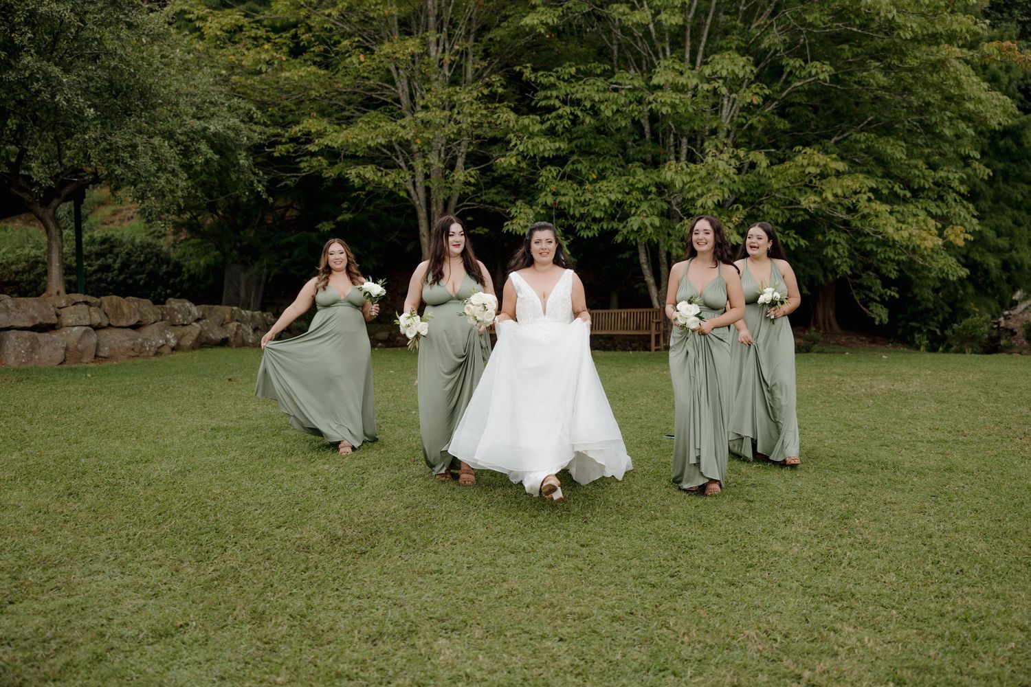 Bride in white gown walks with four bridesmaids in sage green dresses across a lush green lawn.