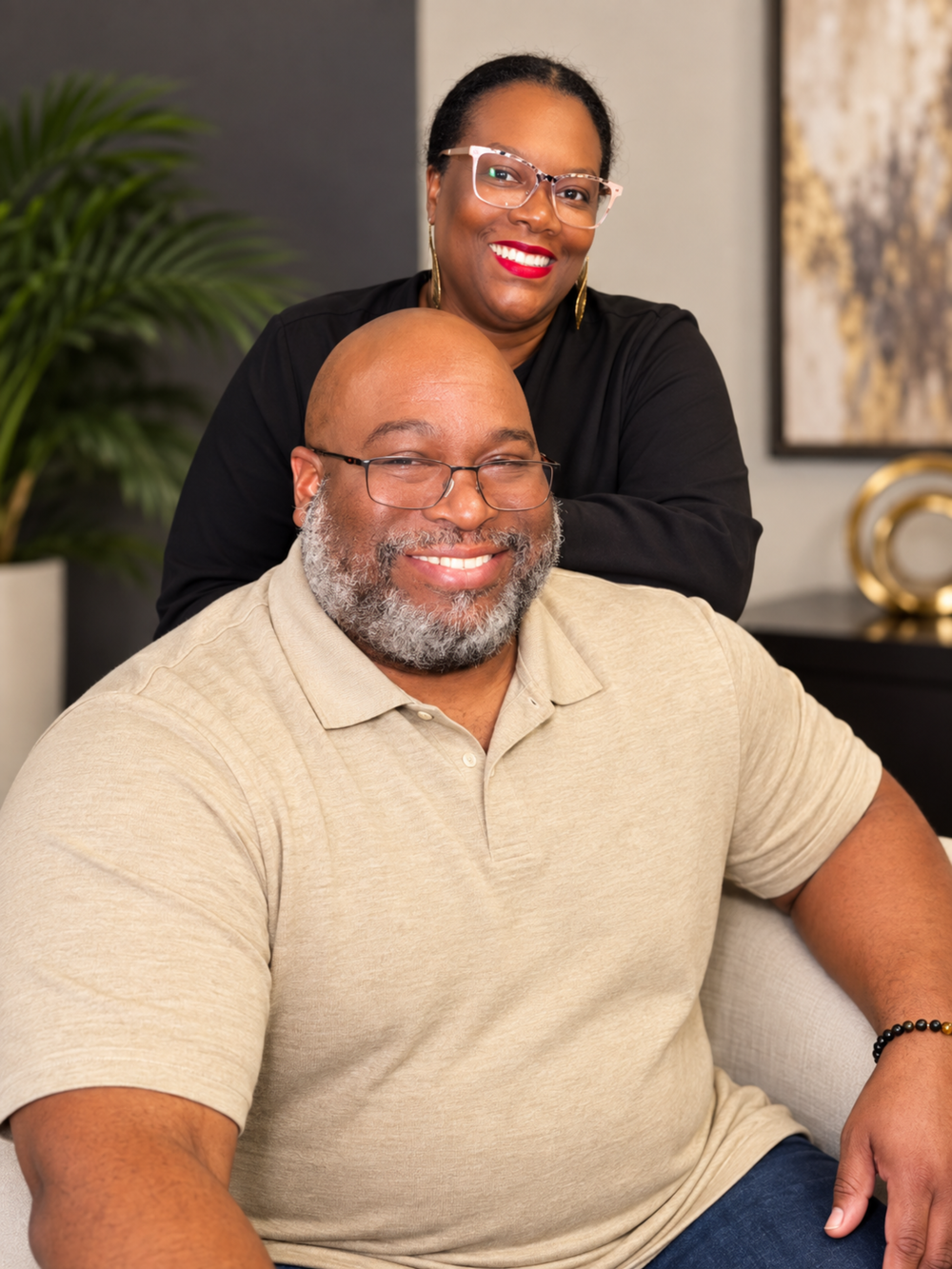 A smiling man with a beard sits as a woman with glasses and red lips stands behind him smiling.