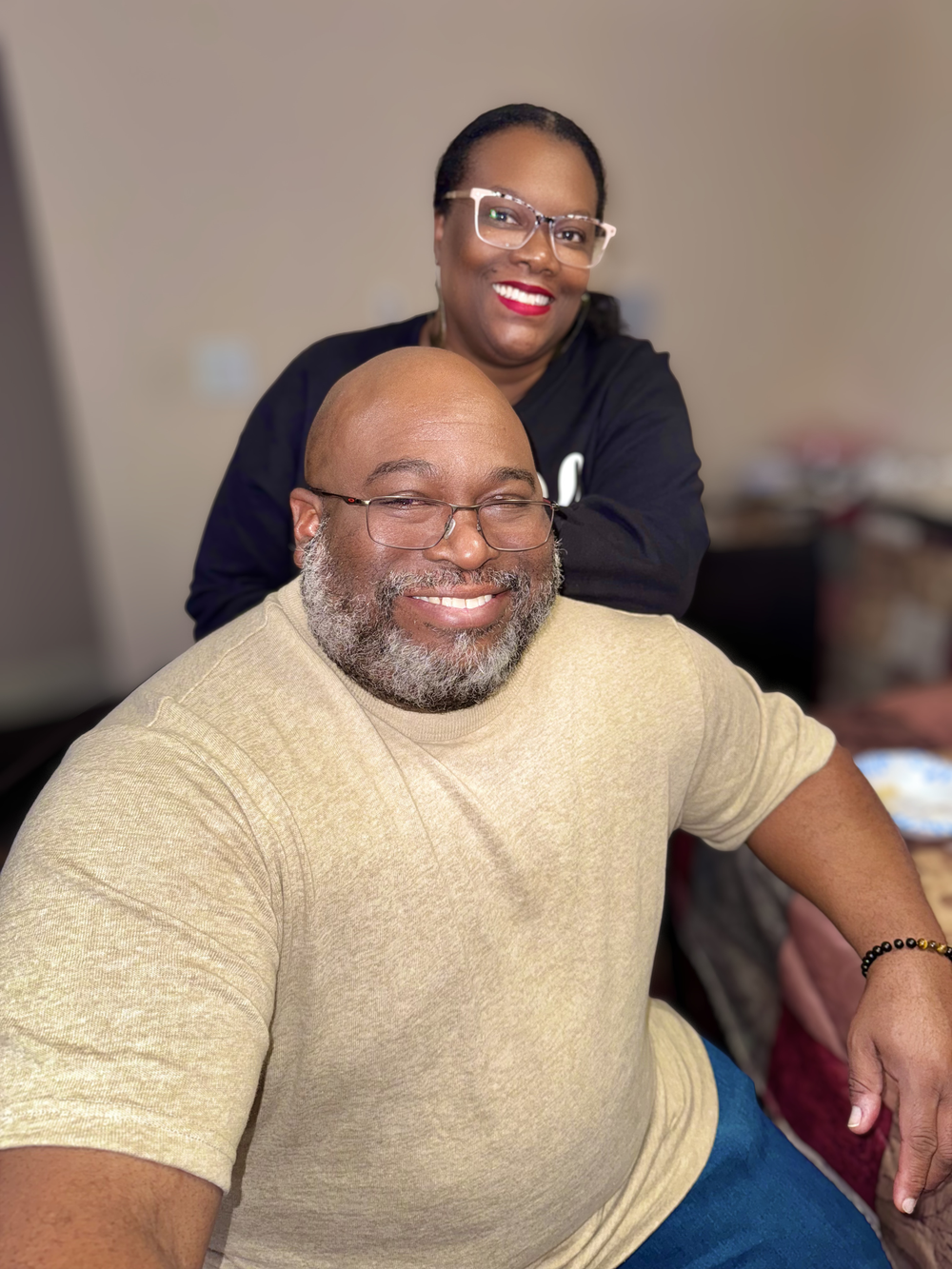 A smiling man with a beard sits as a woman with glasses and red lips stands behind him smiling.