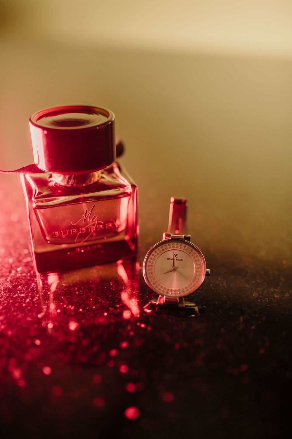 My Burberry perfume bottle beside a delicate rose gold watch on glittery red surface.