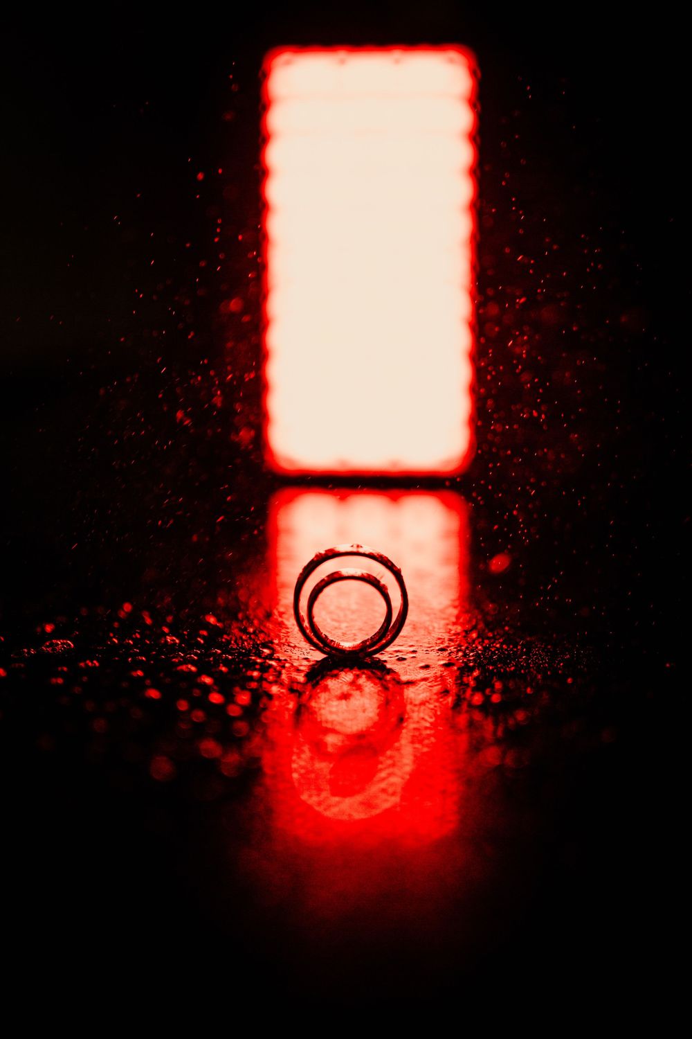 Wedding ring reflected on wet surface with glowing red light in dark background.
