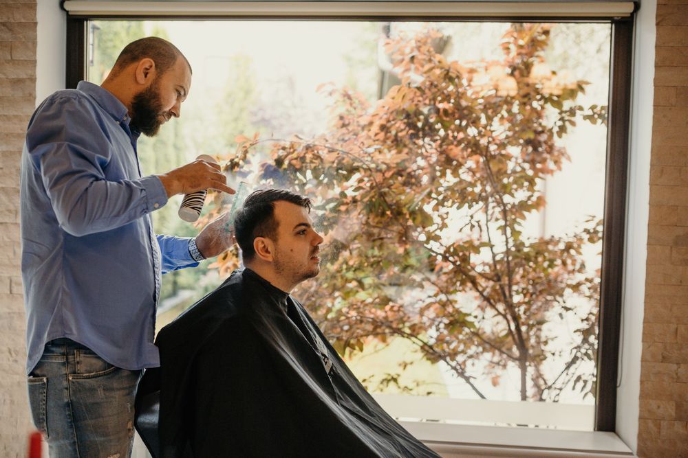 Professional barber spraying water on client's hair near large window with autumn trees.