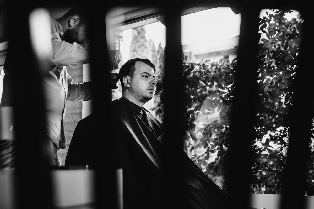 Black and white portrait of groom getting haircut framed through wooden slats.