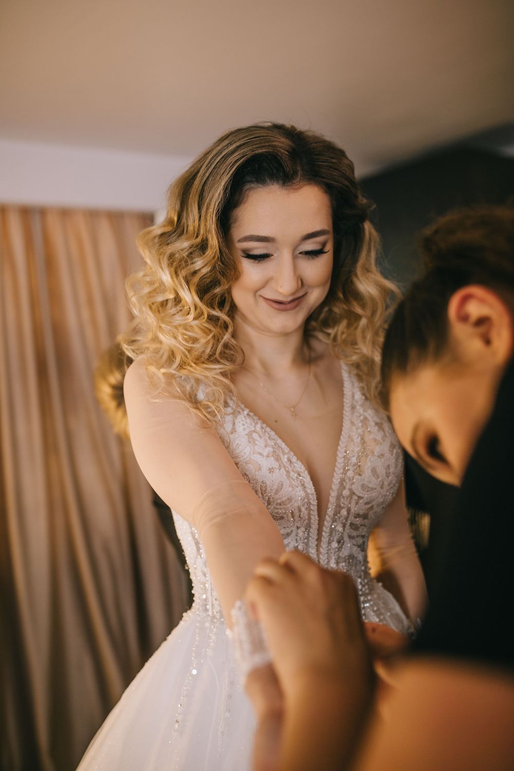 Smiling bride in lace wedding dress being helped to dress by bridesmaid.