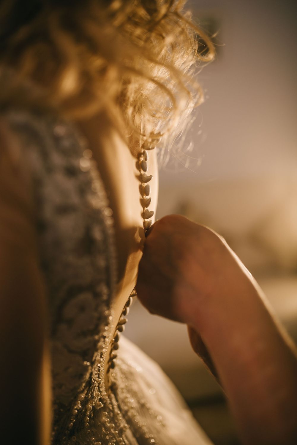 Close-up of hands fastening pearl buttons on back of lace wedding dress.