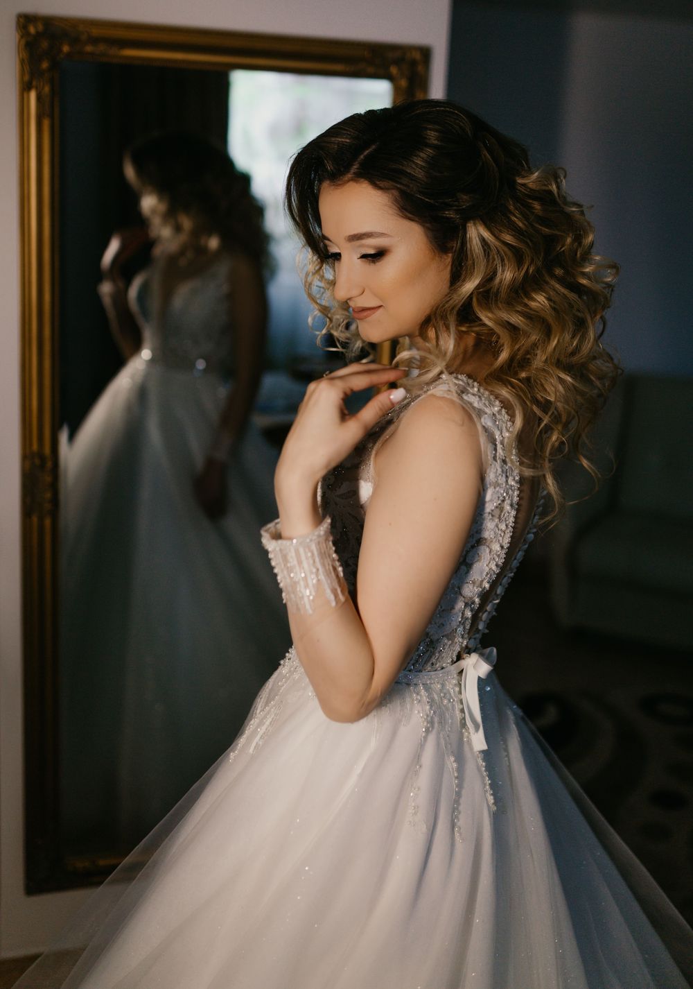 Bride in a beaded ballgown with long curly hair posing beside a gold-framed mirror.