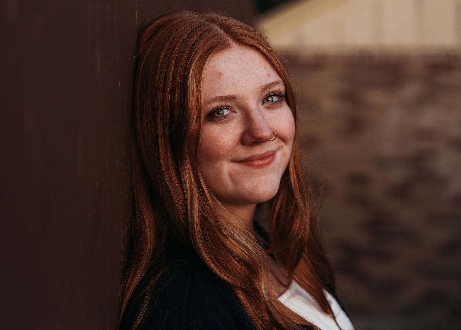 Young woman with long red hair smiling warmly, leaning against a rustic wall in a professional portrait.