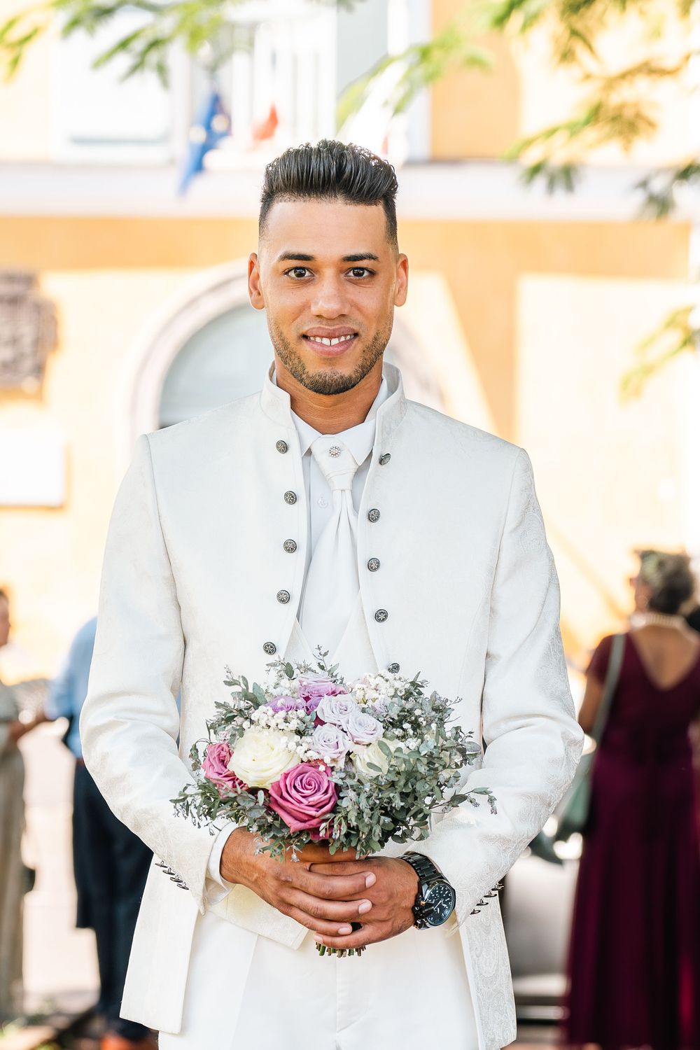 Young groom in white suit holding pink and white flower bouquet outdoors at wedding ceremony.