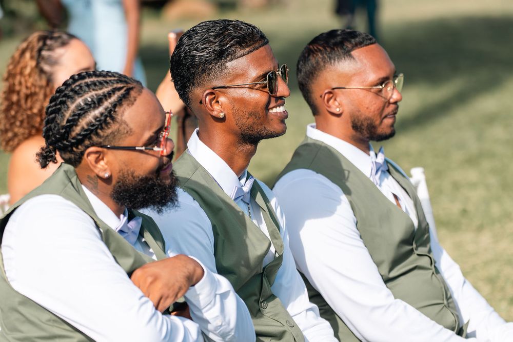 Three groomsmen in sage vests and white shirts smile during an outdoor wedding ceremony.