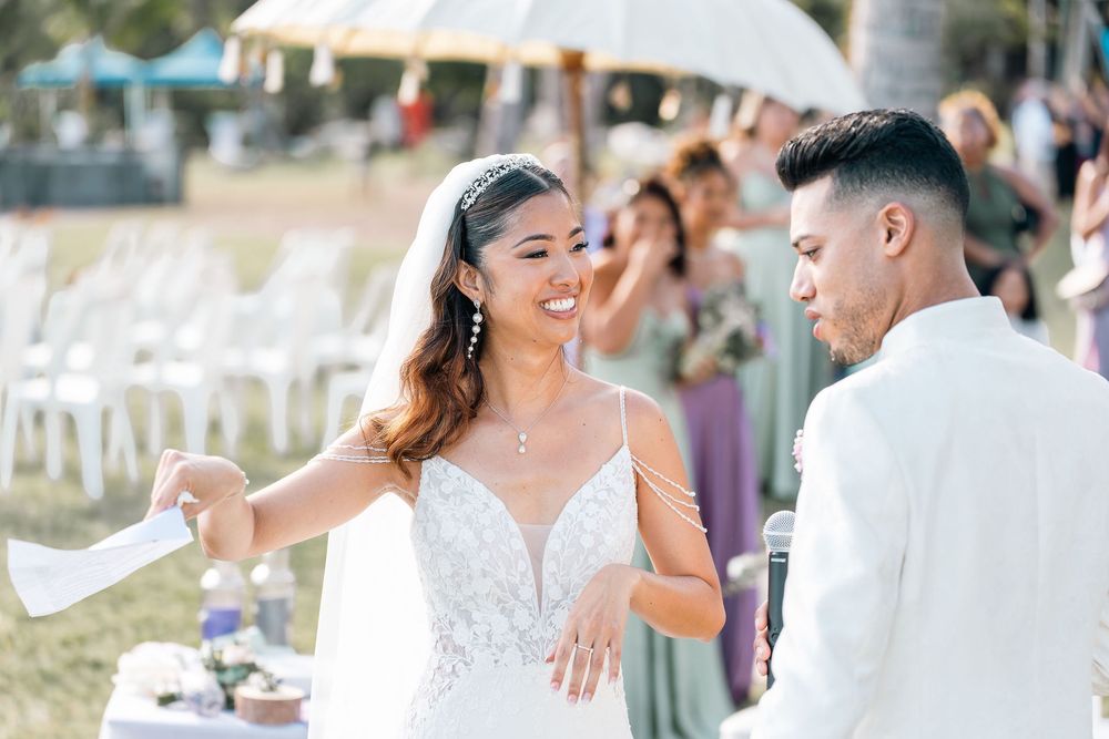 Bride in lace wedding gown smiling joyfully at groom during outdoor wedding ceremony with guests seated in background.